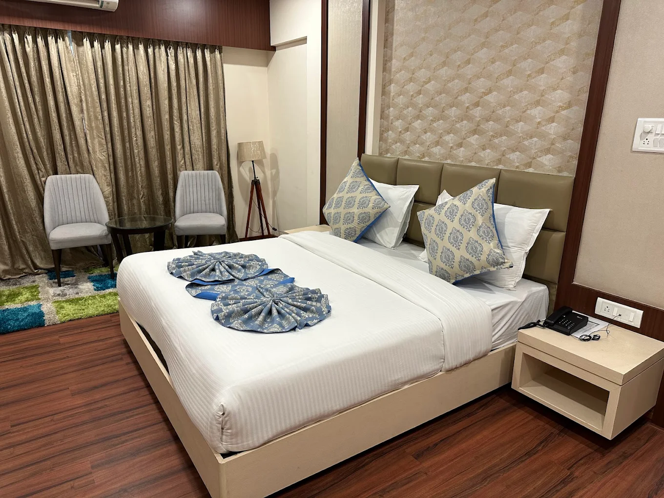 A photograph of a Hotel Swamiraj Deluxe Room, showcasing a king-size bed, elegant furnishings, and a view of the city skyline through large windows. The room is well-lit and inviting.